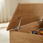 A tampa metálica de segurança mantém aberta a pesada tampa de madeira da caixa de brinquedos enquanto a criança a coloca no interior.